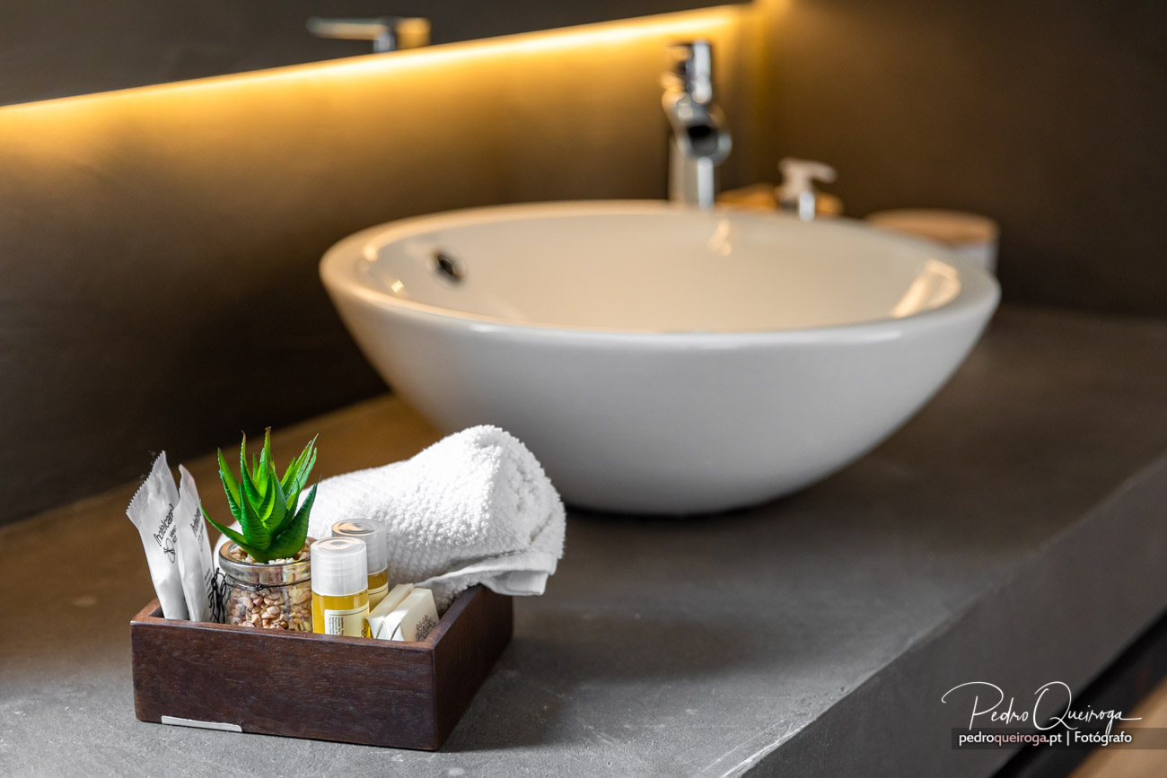 Lavatório branco oval sobre bancada cinza com caixa de amenities e toalha branca em primeiro plano