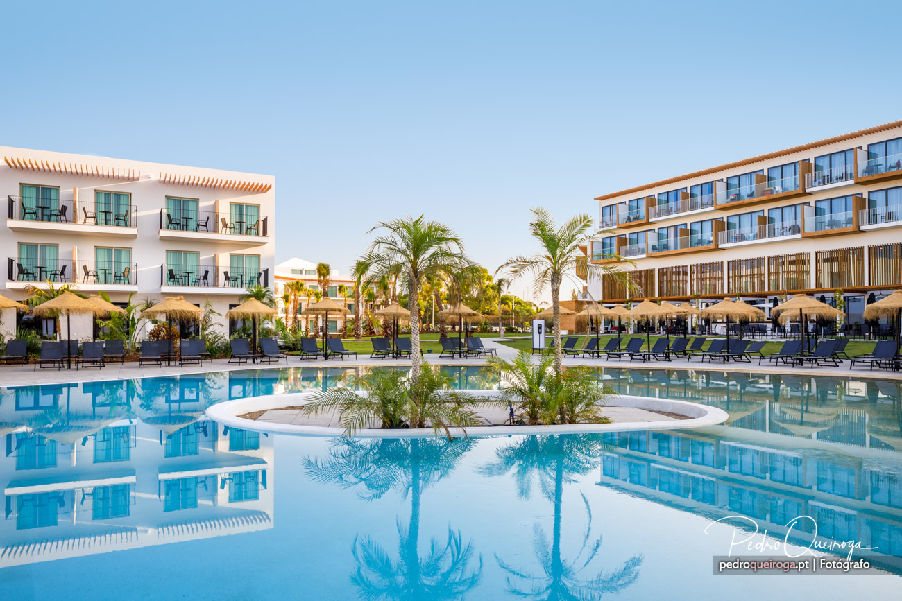 Piscina ampla de hotel de luxo rodeada por palmeiras, espreguiçadeiras e edifícios modernos sob céu azul claro