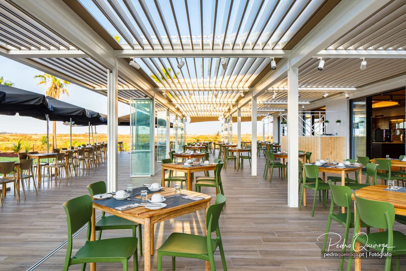 Esplanada moderna com pérgula bioclimática, mesas em madeira, cadeiras verdes e vista para paisagem dourada ao entardecer