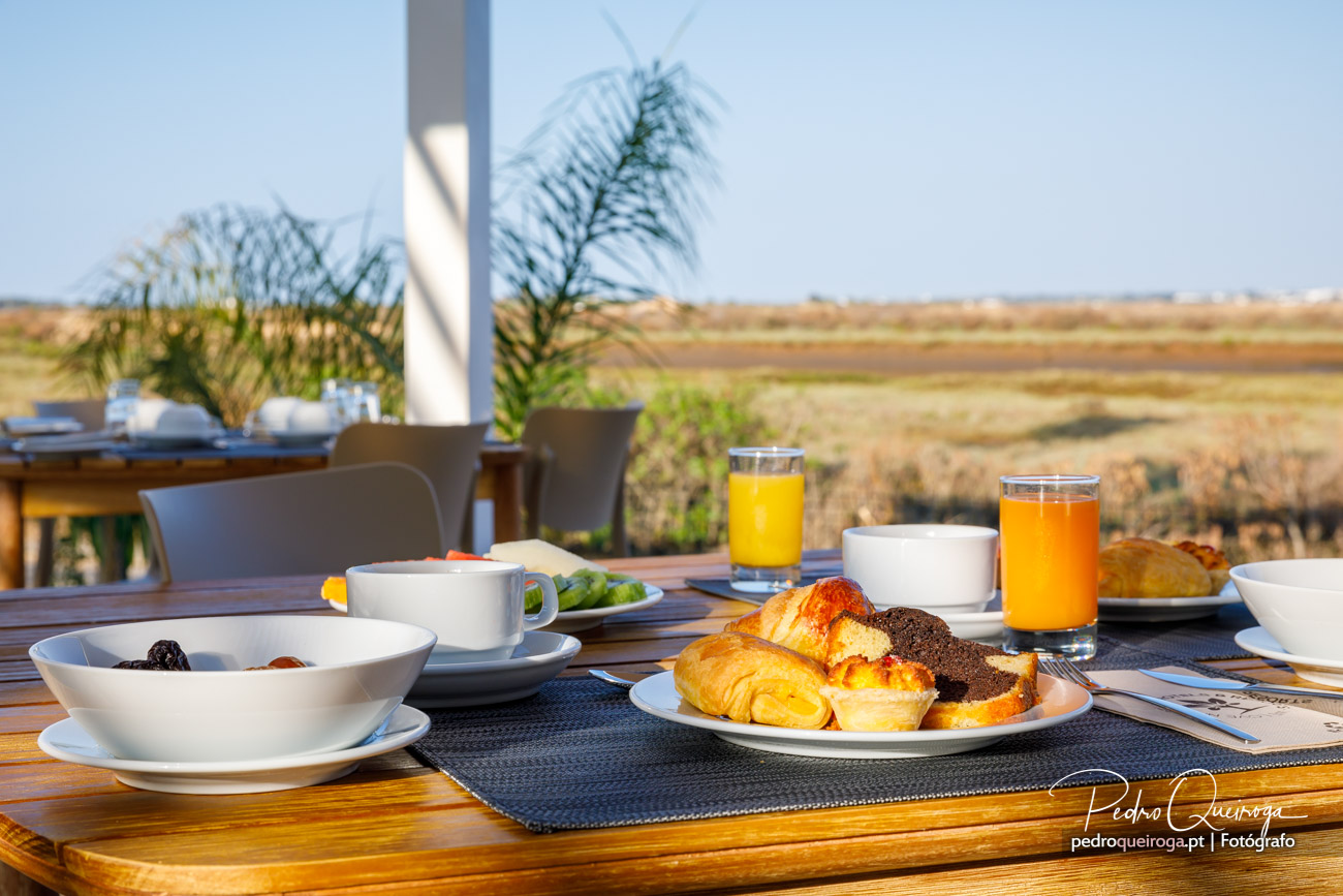 Mesa de pequeno-almoço ao ar livre com croissants, sumos naturais e pastelaria sobre paisagem rural alentejana