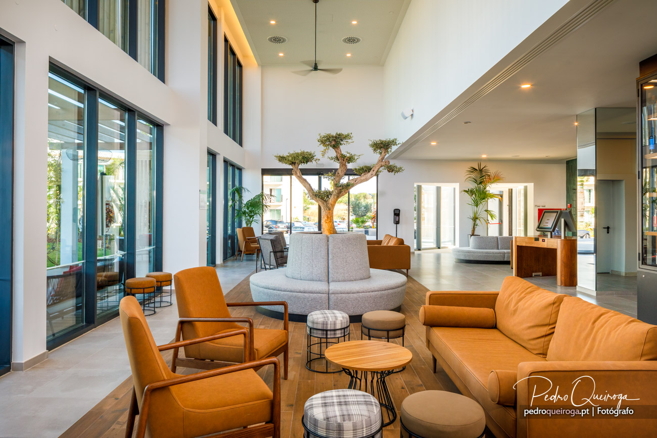 Lounge moderno com pé direito alto, sofás laranja, árvore decorativa central e grandes janelas de vidro a envolver o espaço