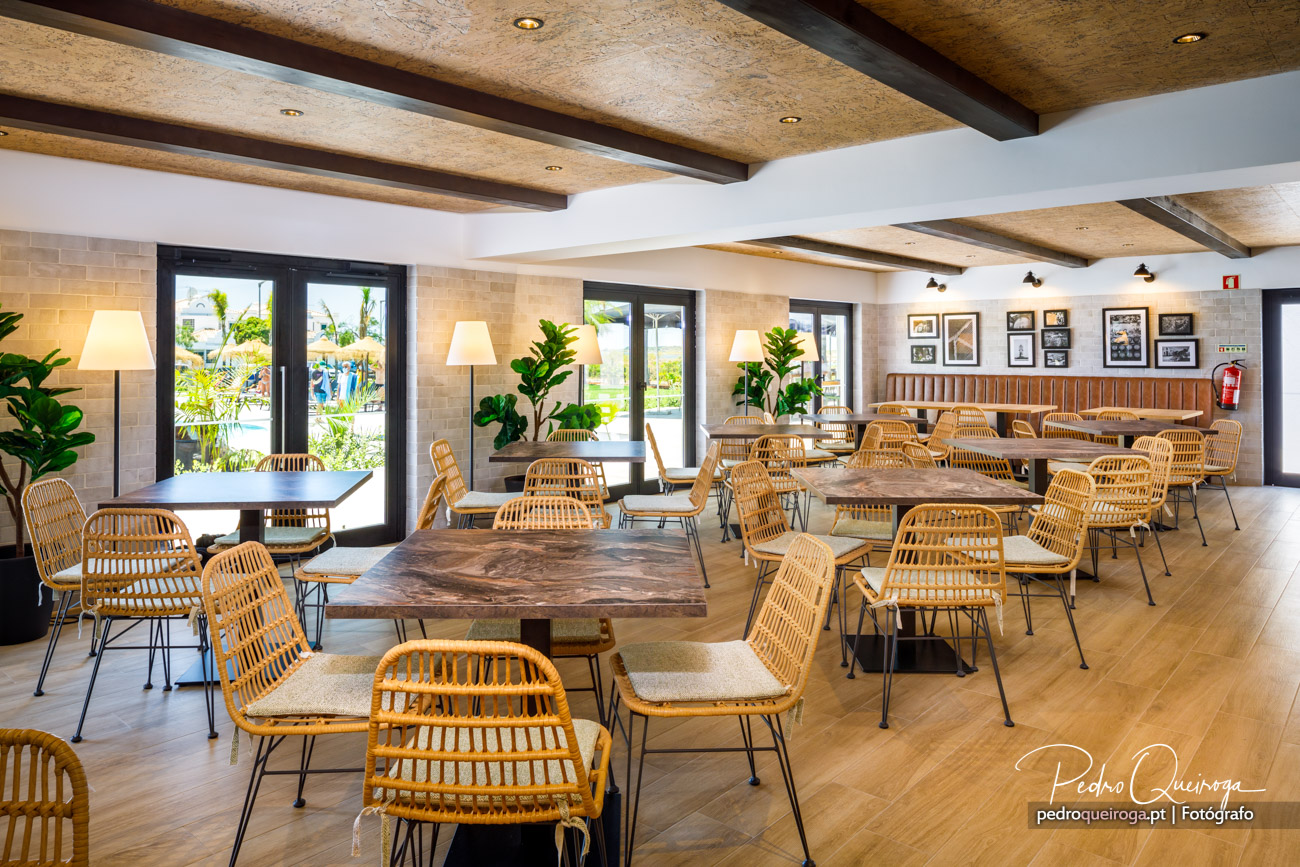 Espaço amplo e luminoso com mesas de madeira rústica, cadeiras em rattan e vigas no teto com iluminação embutida.