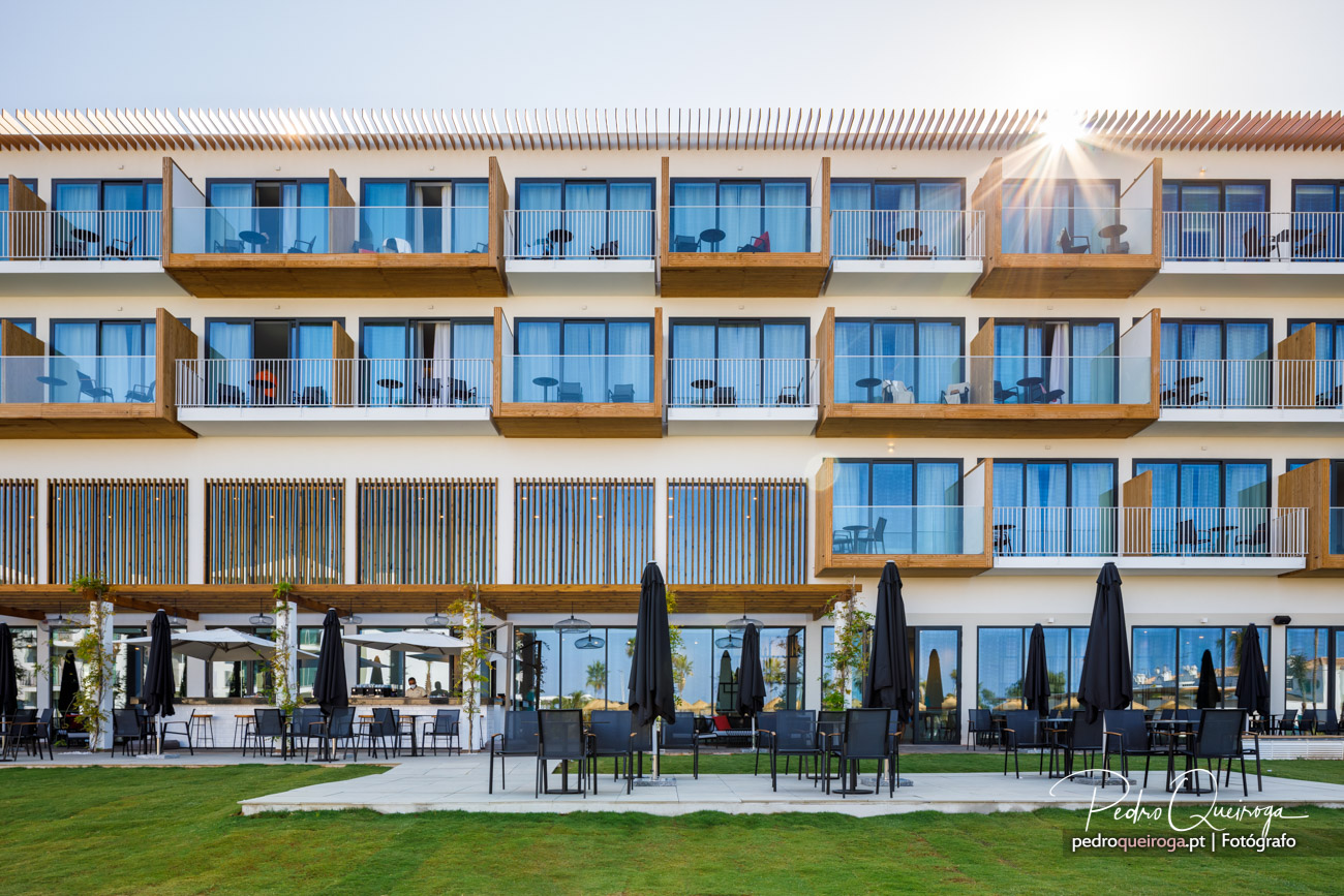Fachada moderna de hotéis com varandas em madeira, esplanada com guarda-sóis e luz solar ao entardecer.