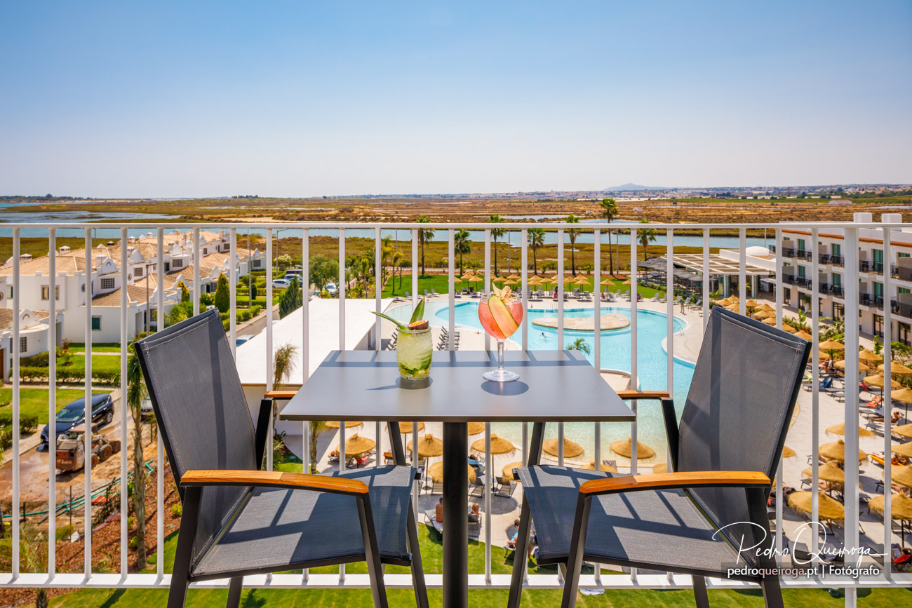 Varanda com mesa, cadeiras e cocktails coloridos, vista panorâmica sobre piscina, resort e zona lagunar ao fundo