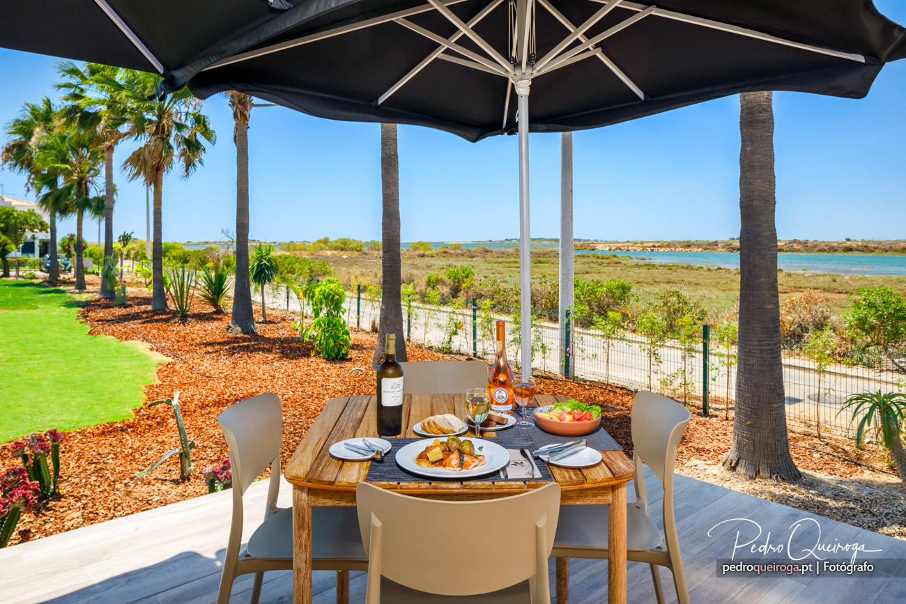 Mesa de refeição ao ar livre sob chapéu de sol, com vista para jardim tropical e paisagem costeira algarvia