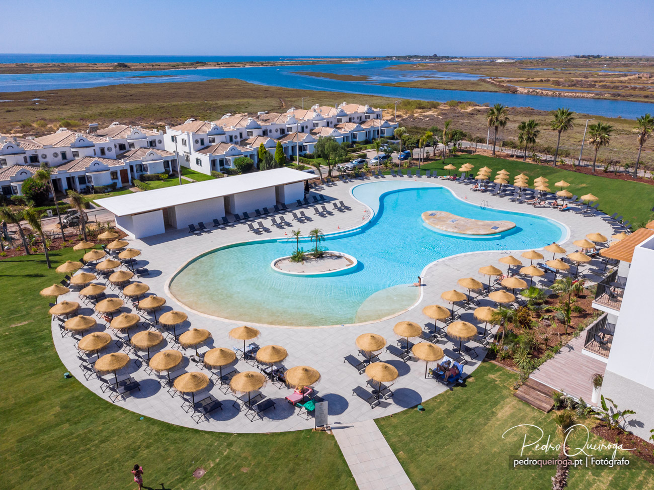Vista aérea de resort com piscina de formas orgânicas, chapéus de palha, moradias brancas e ria ao fundo.