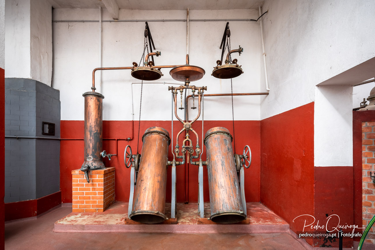 Antigo alambique de cobre com dois cilindros verticais e tubagens suspensas numa sala com paredes vermelhas e brancas