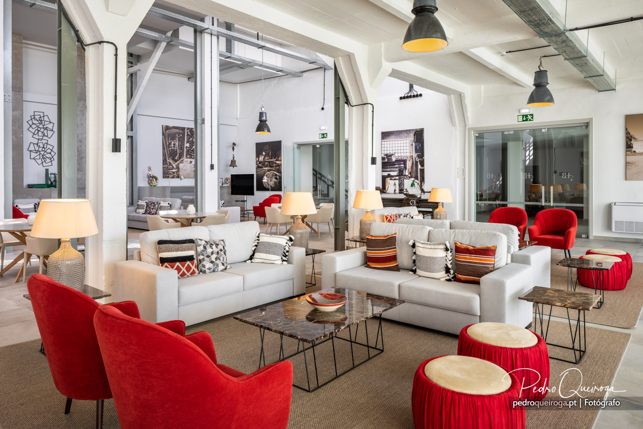 Espaço lounge amplo e luminoso com sofás brancos, poltronas vermelhas, almofadões decorativos e iluminação industrial moderna.