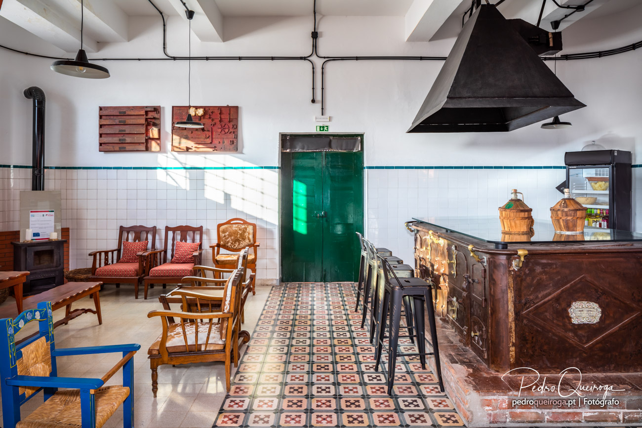 Interior rústico com balcão antigo em madeira, cadeiras vintage, azulejos brancos e porta verde ao fundo.