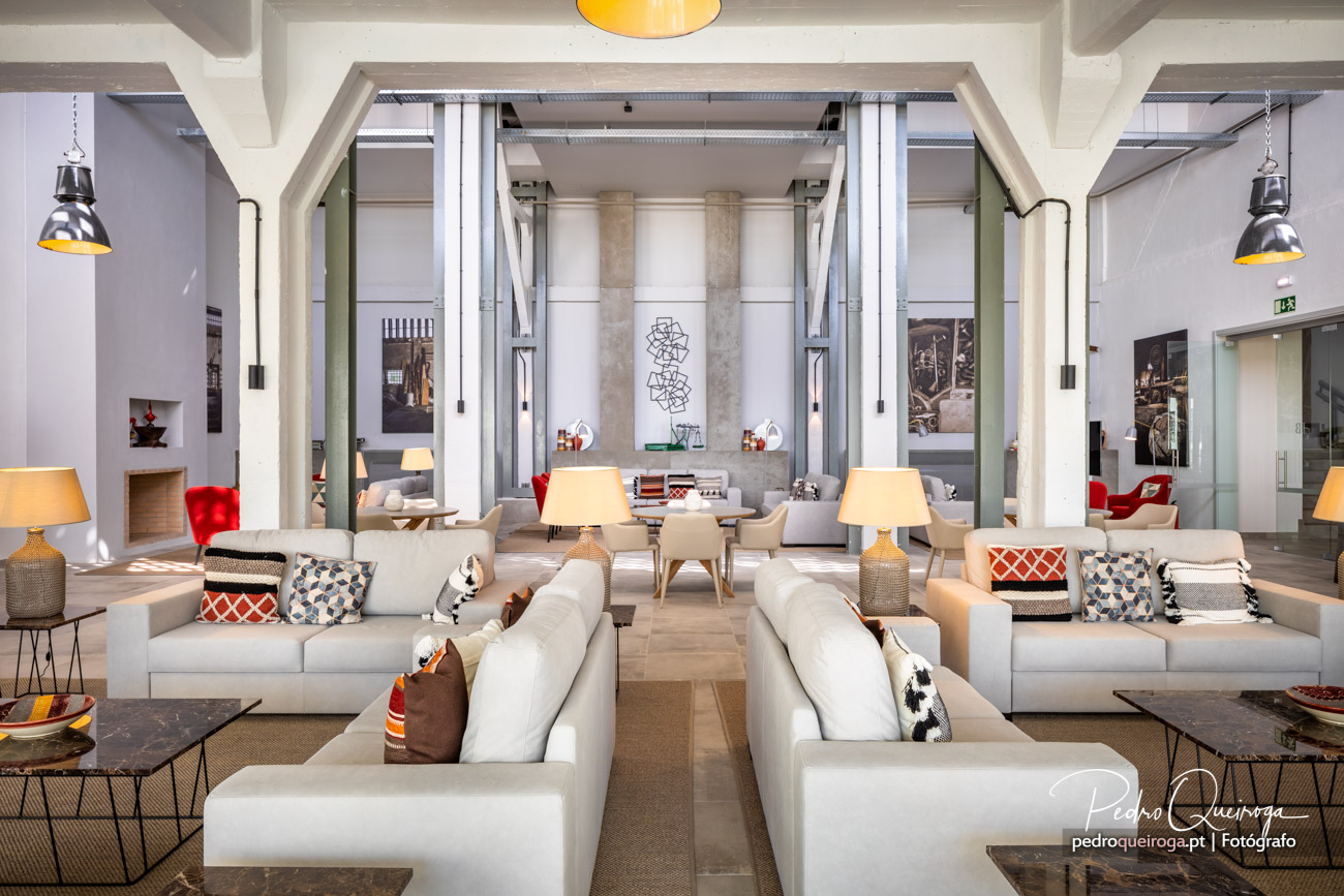 Salão amplo e luminoso com sofás brancos, almofadas coloridas, candeeiros pendentes e pilares brancos de estilo industrial