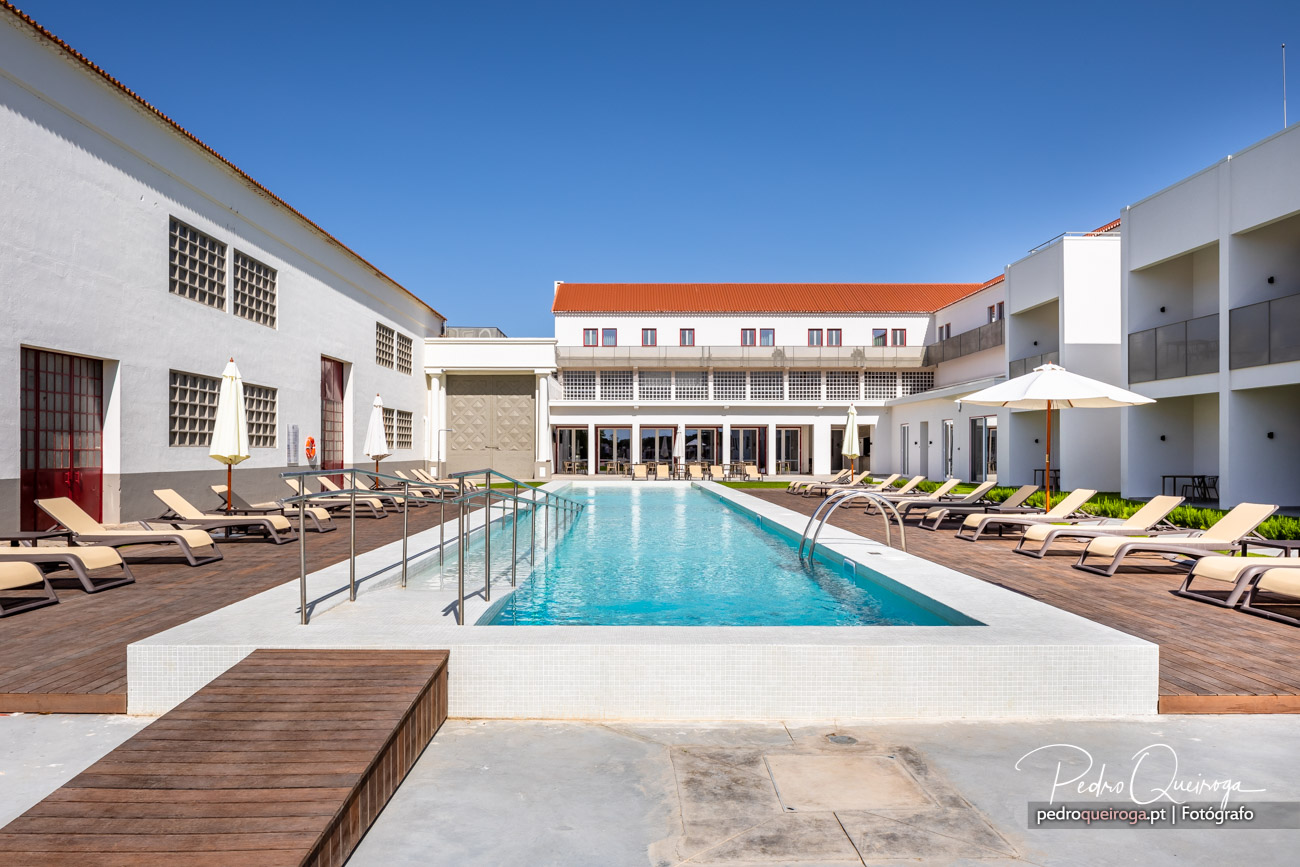 Piscina exterior rodeada por espreguiçadeiras e edifícios brancos com telhado laranja sob céu azul limpo