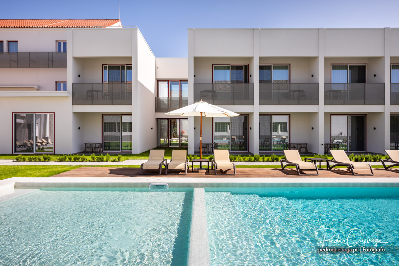 Fachada moderna de apartamentos brancos com varandas, piscina azul cristalina e espreguiçadeiras junto ao relvado.