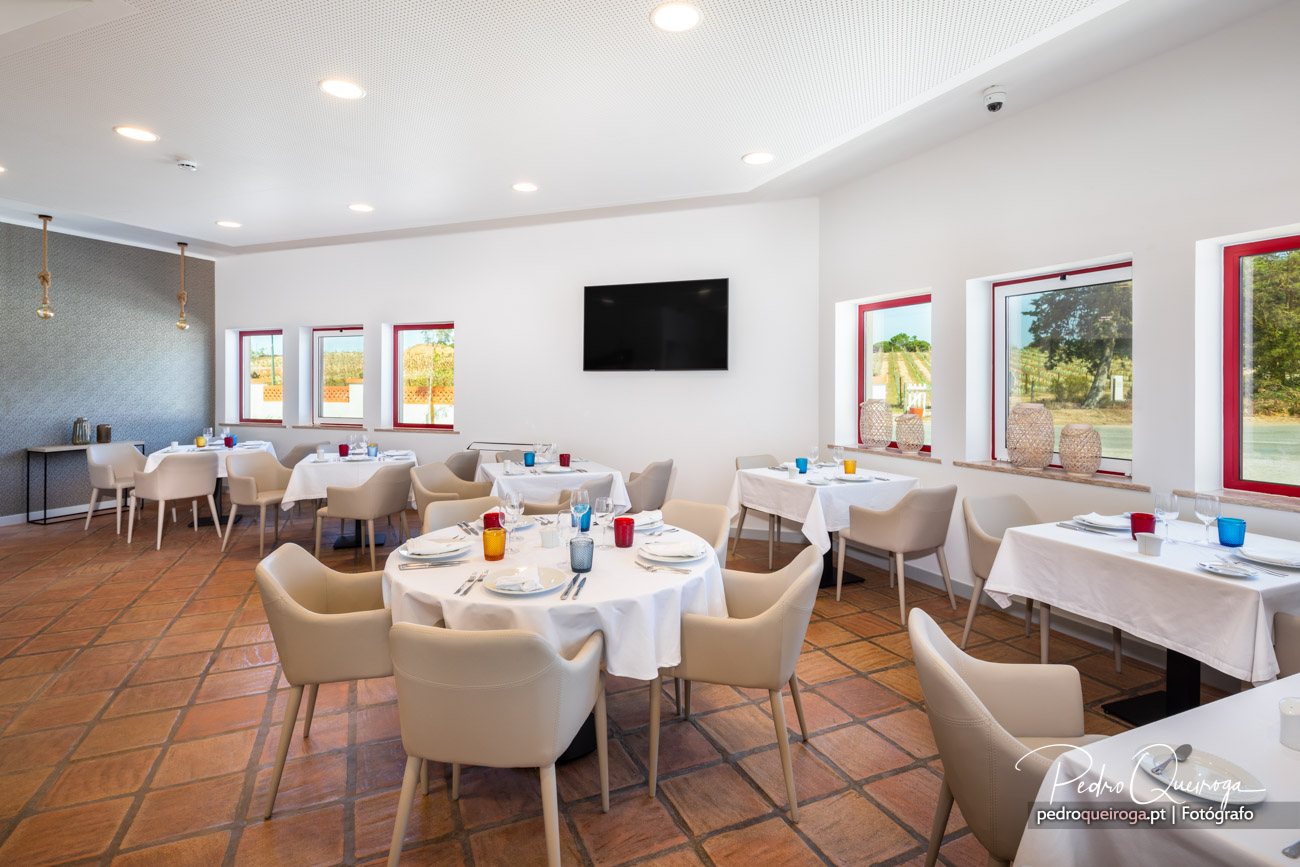 Sala de refeições luminosa com mesas redondas e cadeiras bege, janelas com caixilharia vermelha e vista para o exterior