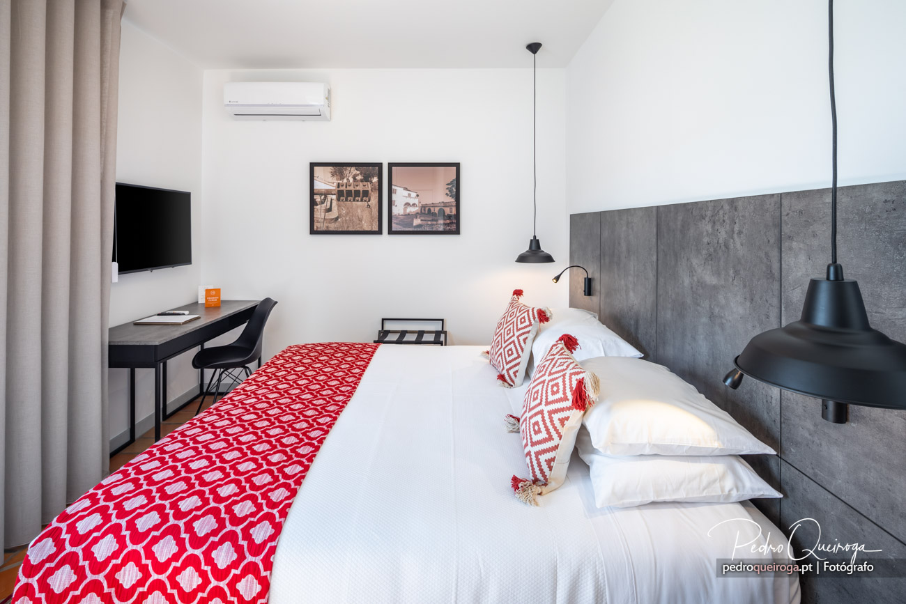 Quarto moderno com cabeceira em betão, roupa de cama vermelha e branca, secretária e candeeiros suspensos pretos.