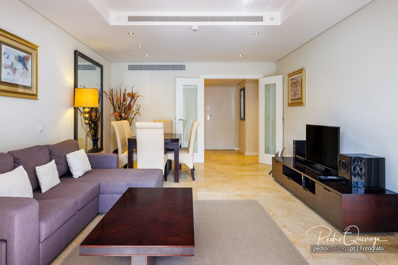 Sala ampla e luminosa com sofá cinza, mesa de jantar, móvel de TV e corredor ao fundo com porta de entrada.