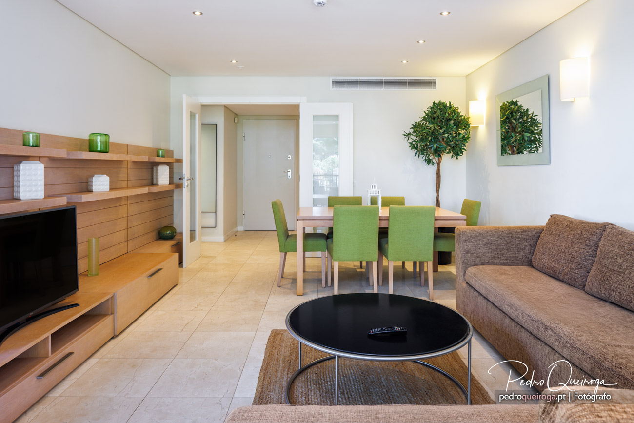 Sala de estar ampla e luminosa com mesa de jantar, cadeiras verdes e árvore decorativa junto à parede branca