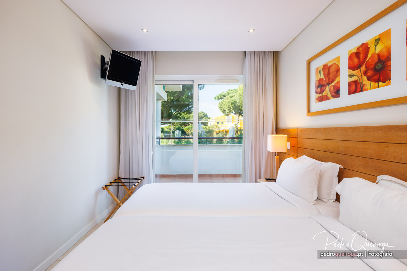 Quarto de hotel luminoso com cama de casal, cabeceira em madeira, televisão na parede e varanda com vista exterior