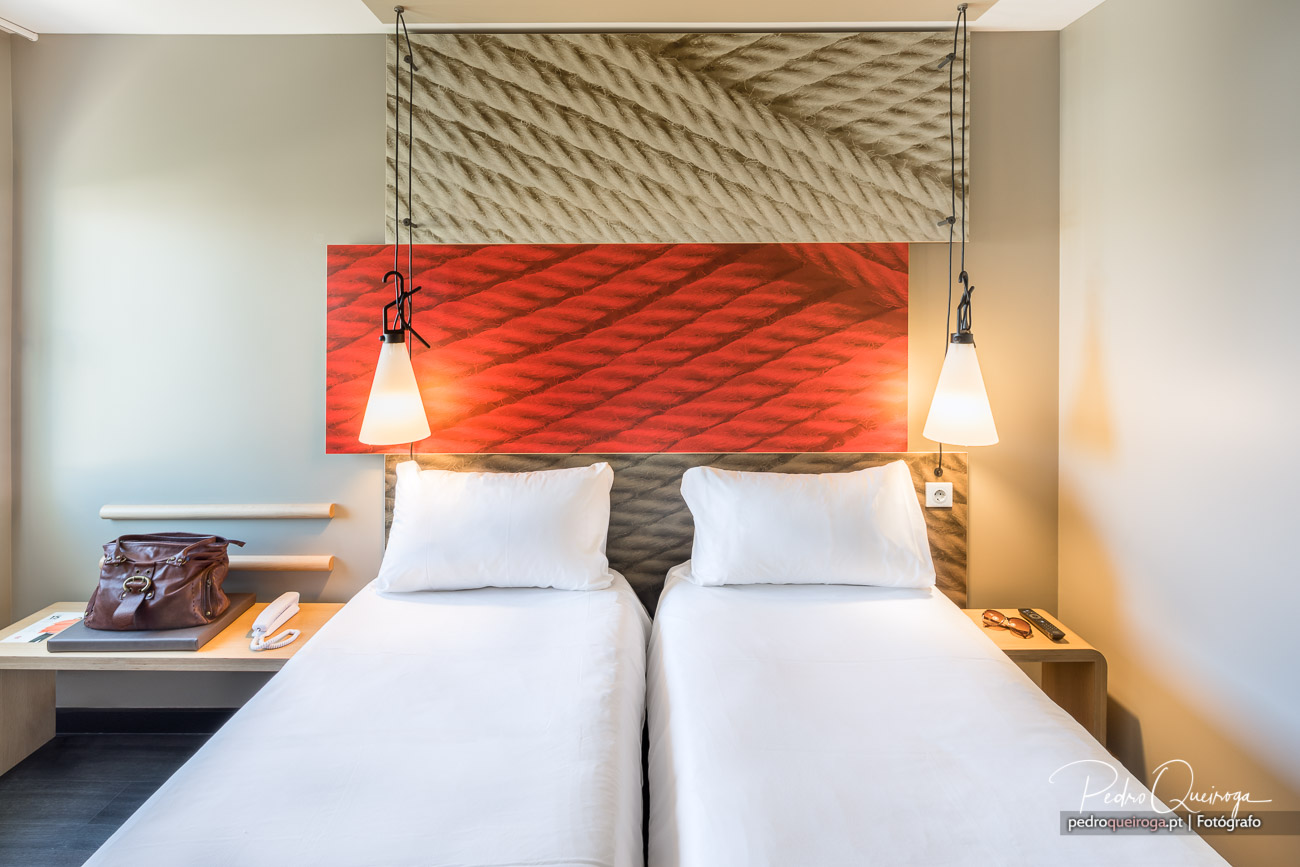 Quarto moderno com duas camas individuais, cabeceira texturizada, painel vermelho vibrante e candeeiros suspensos minimalistas.