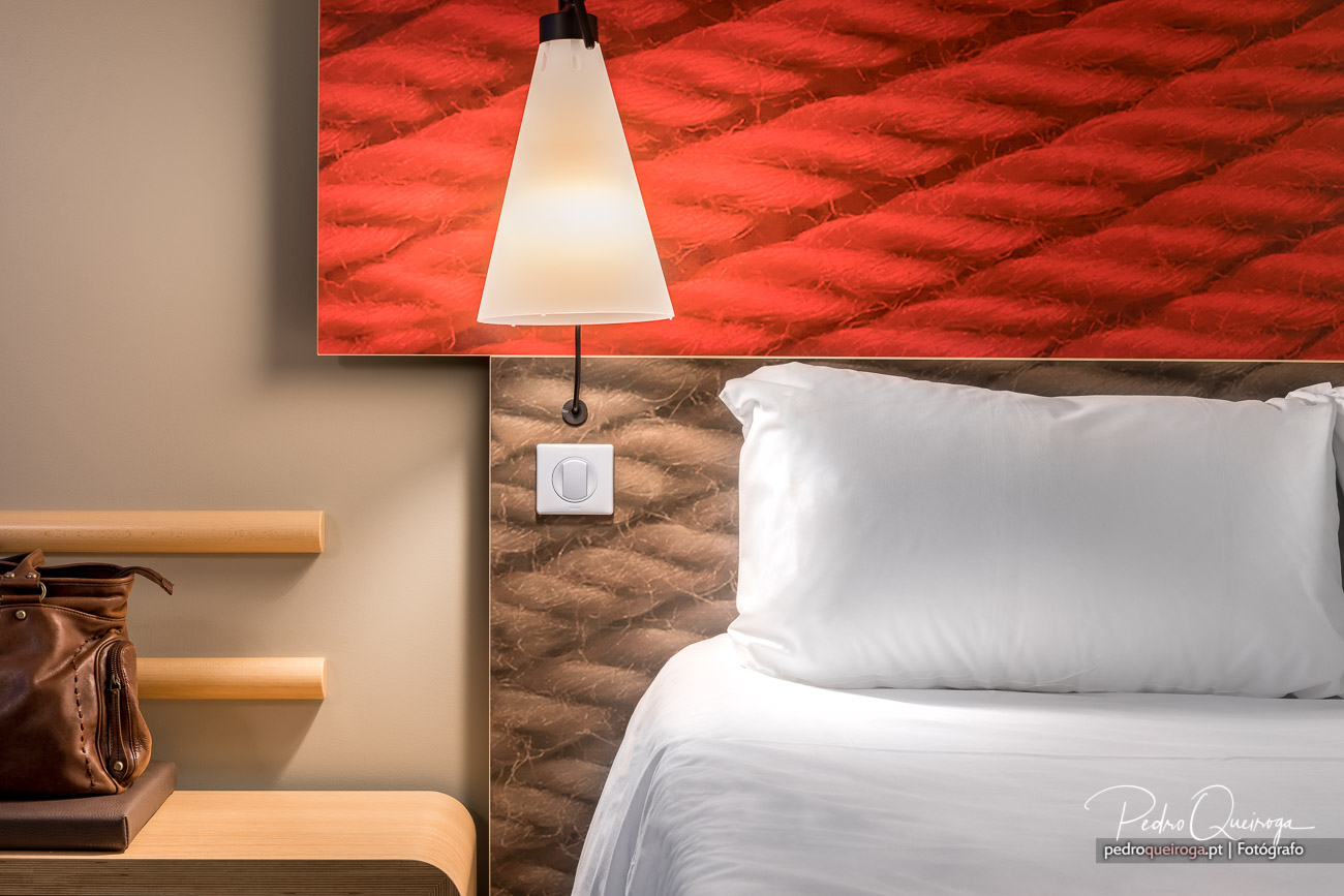 Quarto moderno com cama branca, candeeiro pendente e quadro vermelho vibrante na parede texturizada.