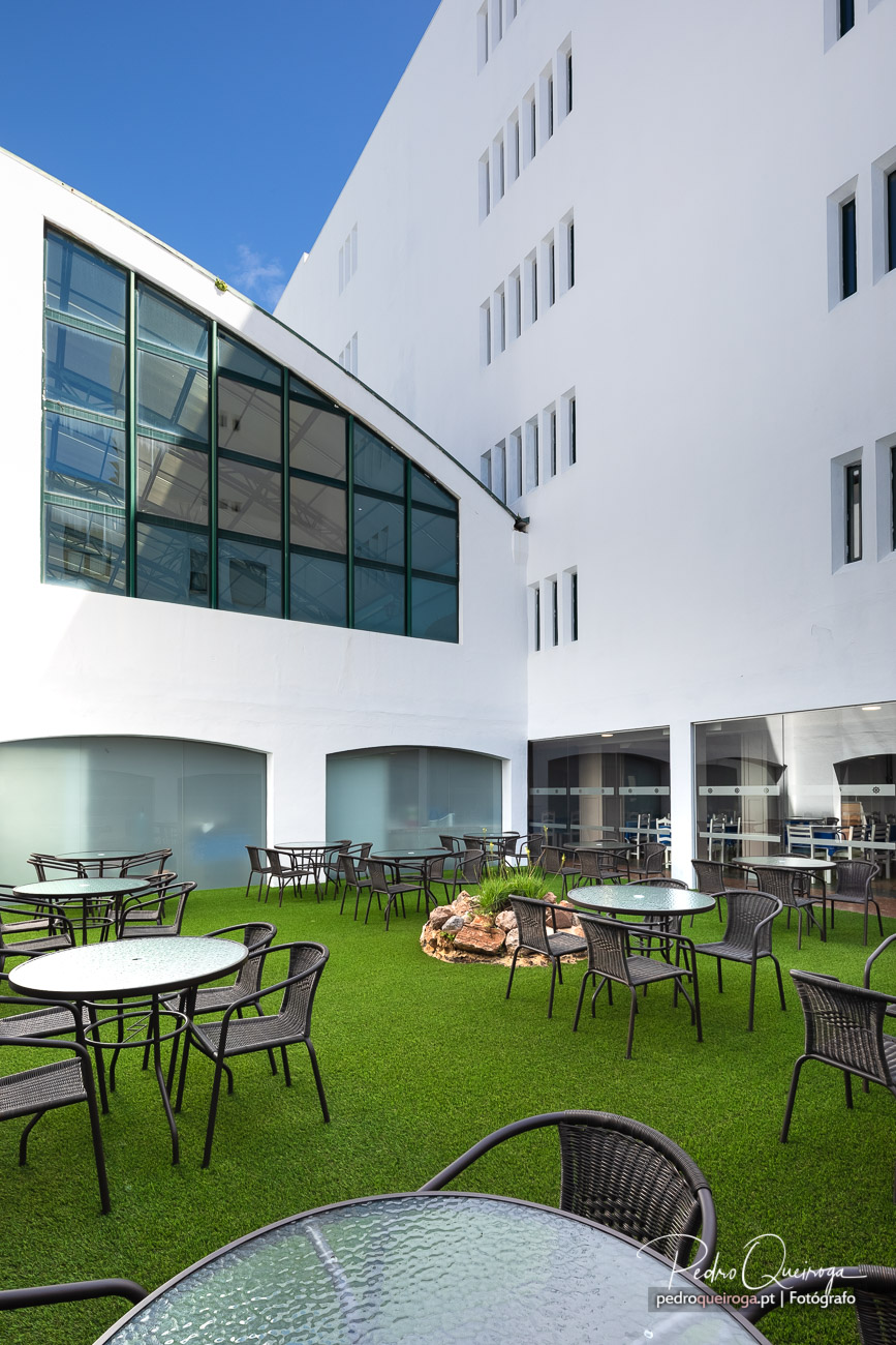 Pátio exterior com esplanada relvada e mesas, rodeado por edifício branco de arquitetura moderna com vidros amplos.