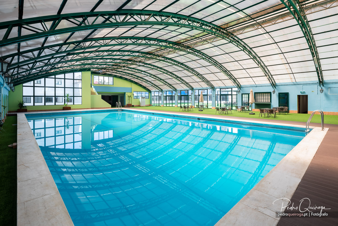 Piscina interior coberta com estrutura metálica verde, paredes coloridas e zona de lazer com mesas ao fundo.