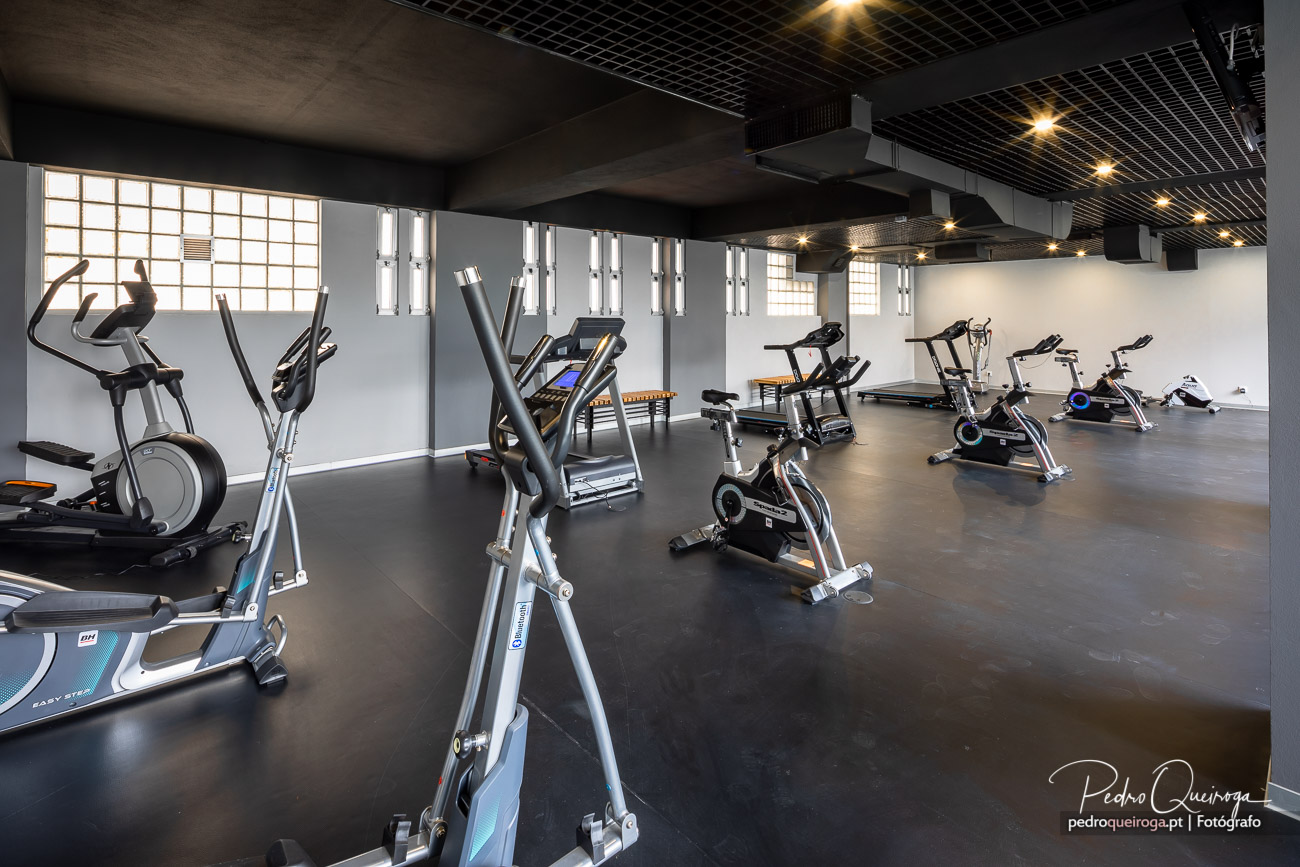 Ginásio moderno e amplo com equipamentos variados, destacando a arquitetura industrial do teto em grelha escura