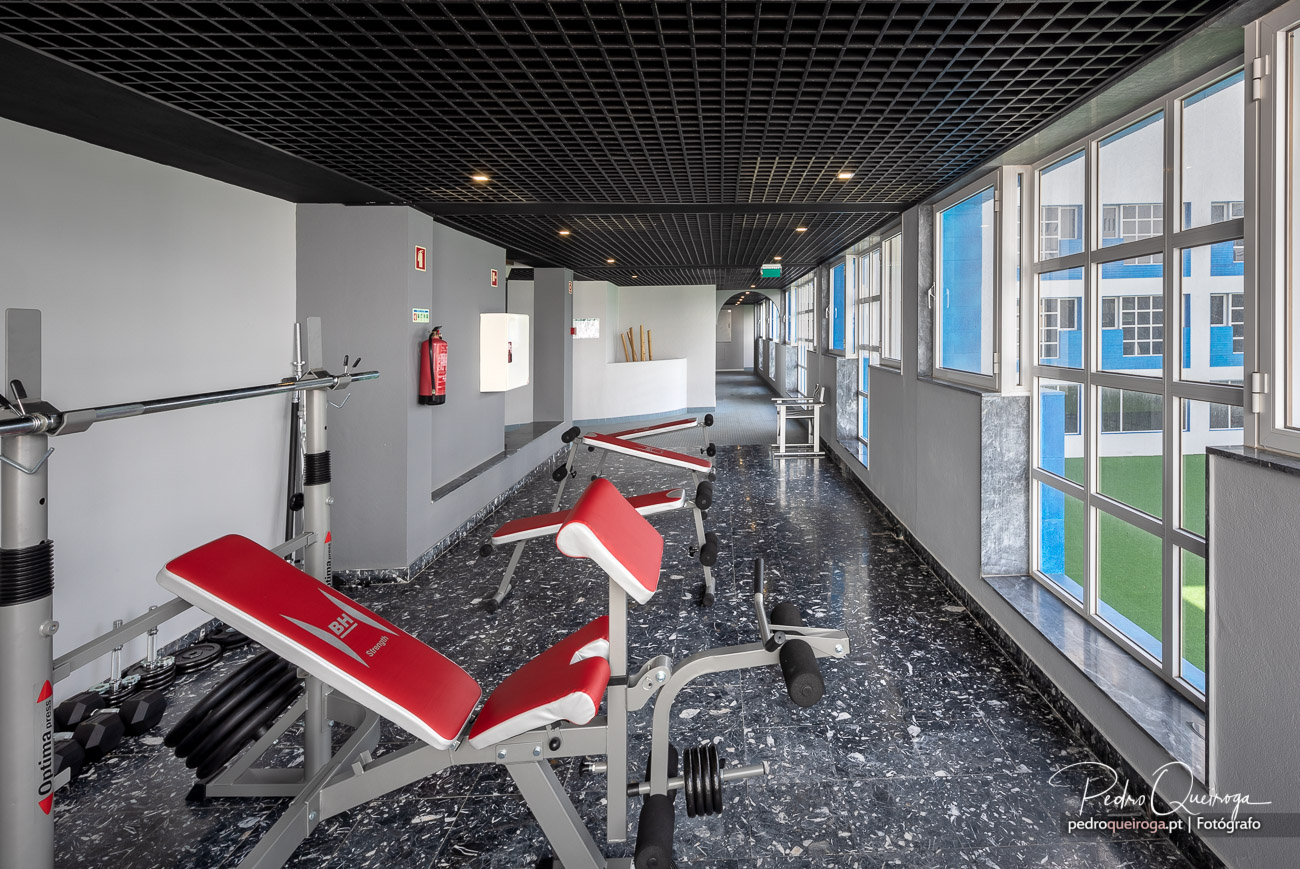 Ginásio moderno com equipamentos vermelhos e brancos, teto negro em grelha e amplas janelas com vista para edifício exterior.
