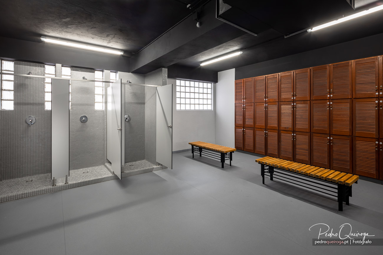 Balneário moderno com chuveiros, cacifos em madeira e bancos, iluminado por luz natural e tubos LED no teto cinza.