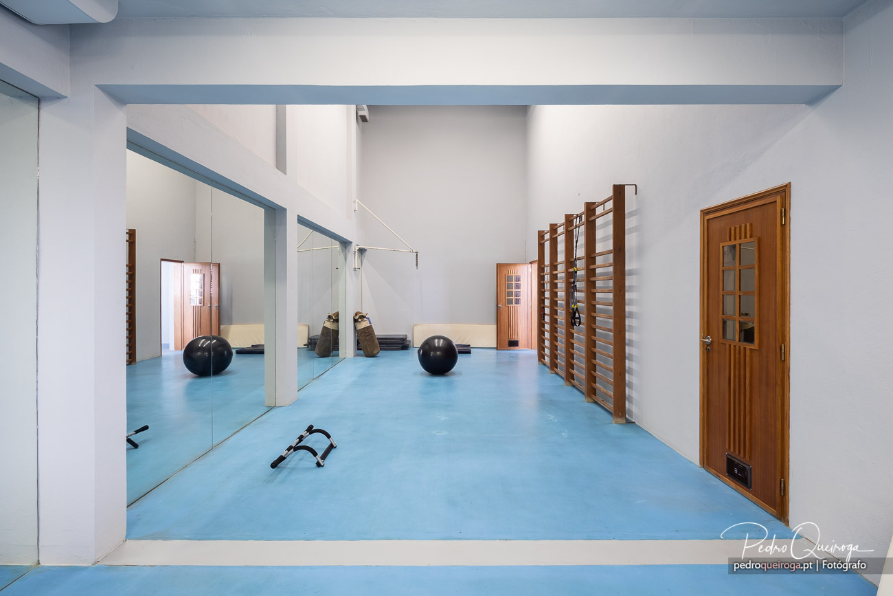 Estúdio de ginástica com piso azul, espelho grande, bola medicinal, escada sueca e porta de madeira.