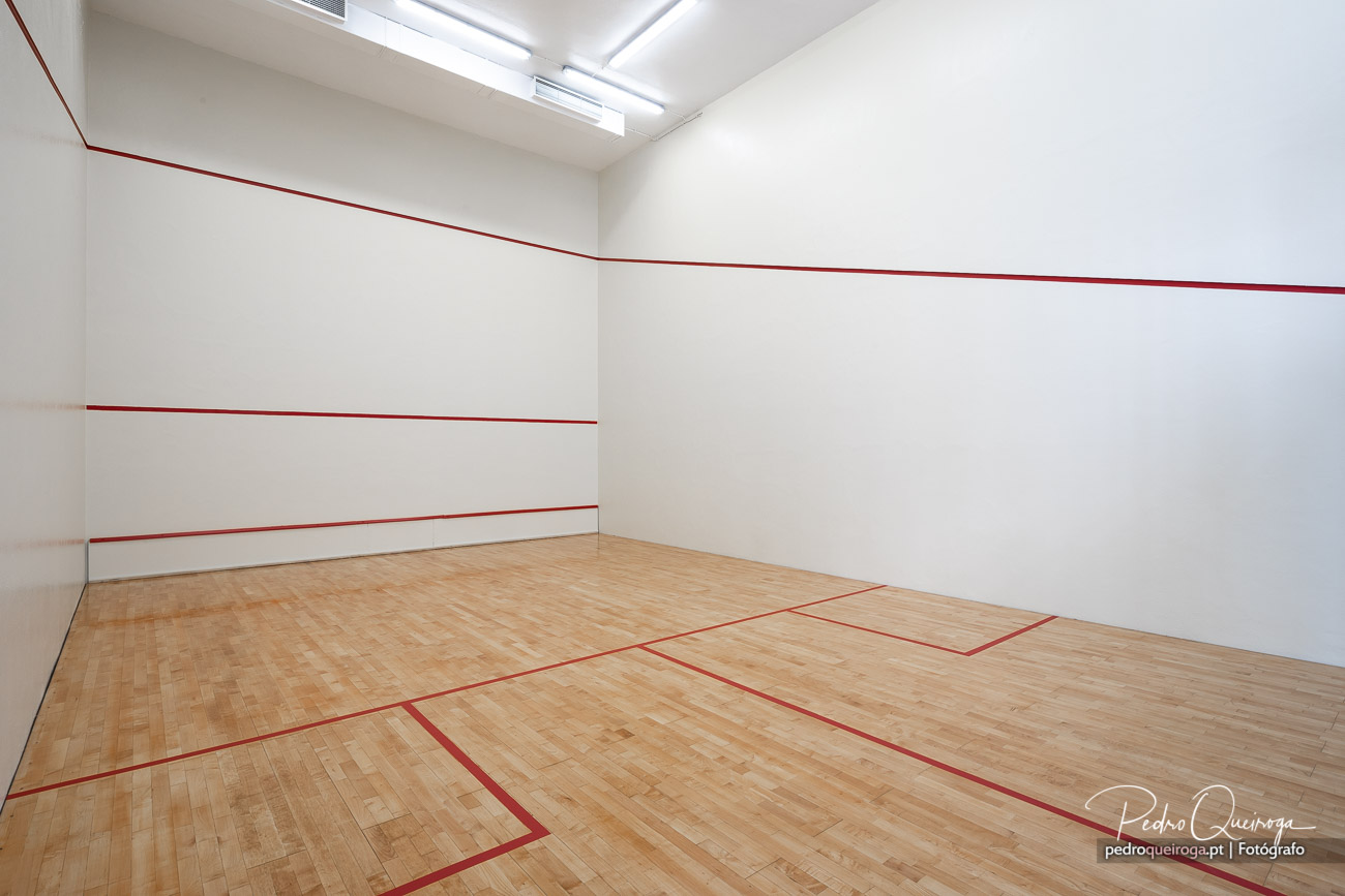 Court de squash com paredes brancas, linhas vermelhas regulamentares e pavimento em madeira clara sob iluminação natural