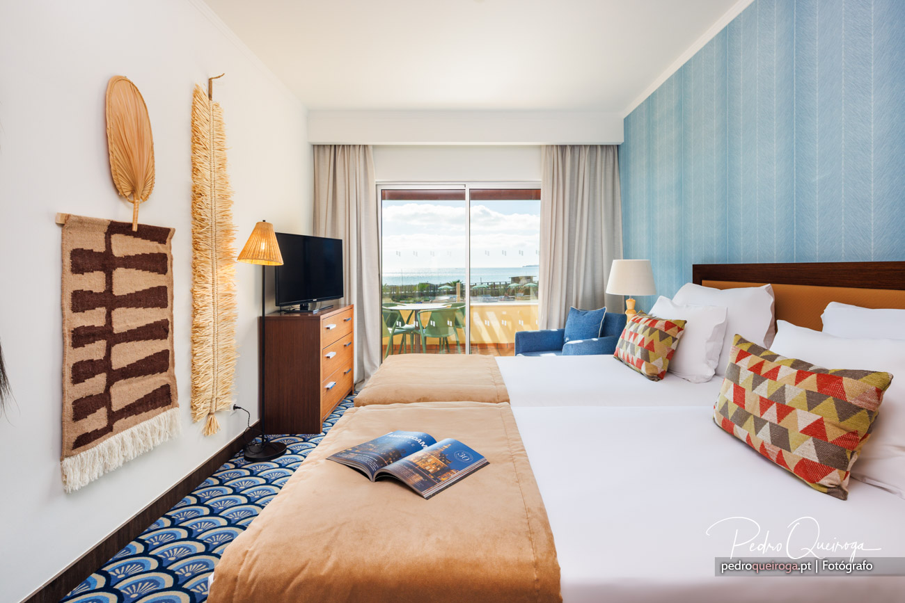 Quarto de hotel luminoso com parede azul, decoração em tons quentes e vista para o mar através da varanda