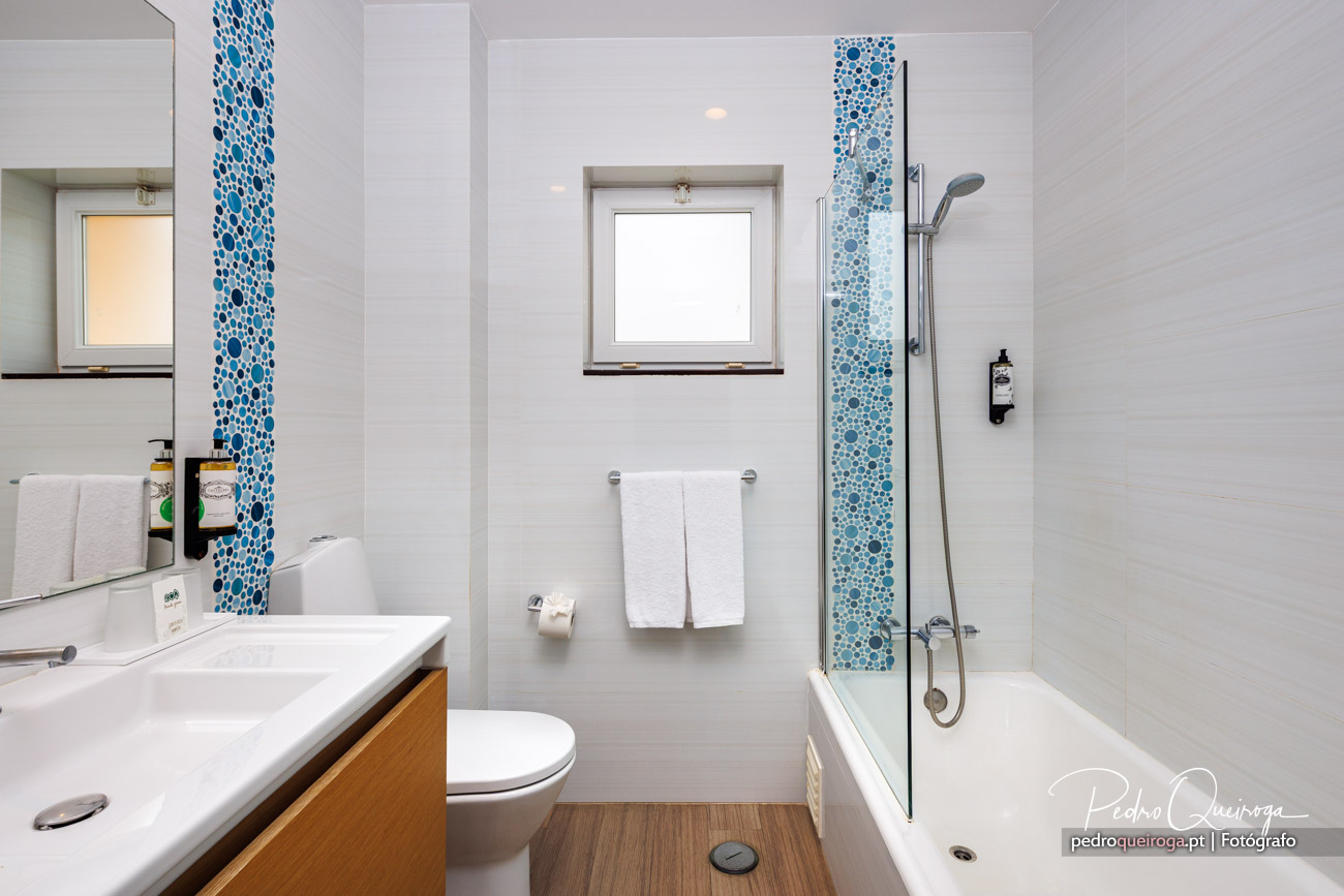 Casa de banho moderna com mosaico azul, banheira com duche, toalheiro branco e móvel de madeira clara