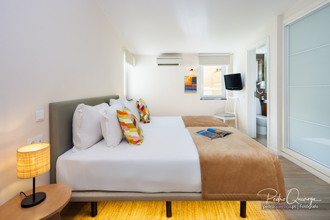 Quarto luminoso com cama de casal, almofadas coloridas, tapete amarelo e casa de banho em suite com porta de vidro