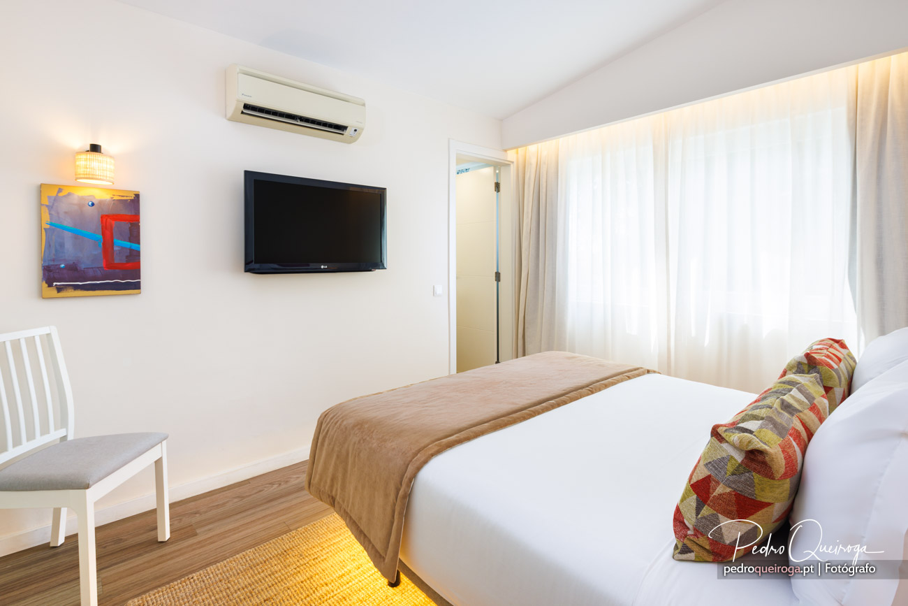 Quarto moderno com cama de casal, televisão na parede, ar condicionado e janelas amplas com cortinas brancas