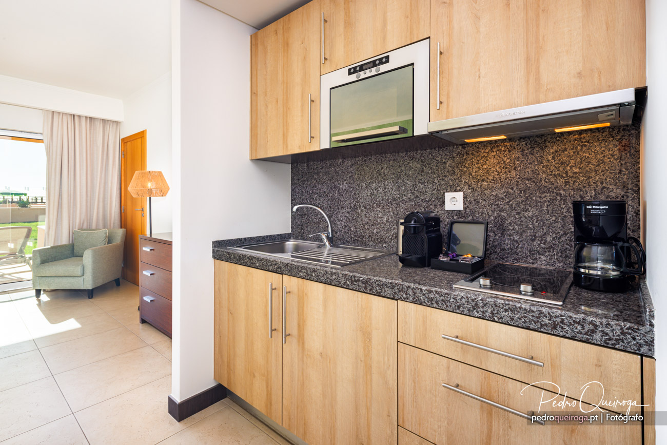 Kitchenette moderna com armários em madeira clara, bancada em granito escuro e pequena sala ao fundo com varanda