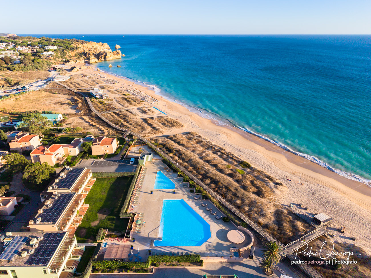 Vista aérea de praia algarvia com piscinas azuis, vegetação dourada, falésias ao fundo e mar turquesa cristalino