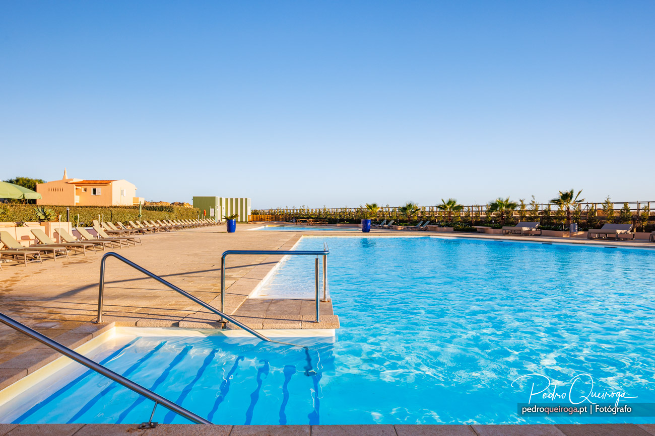 Piscina ampla com água cristalina, espreguiçadeiras bege, palmeiras e céu azul ao fundo num resort ensolarado.