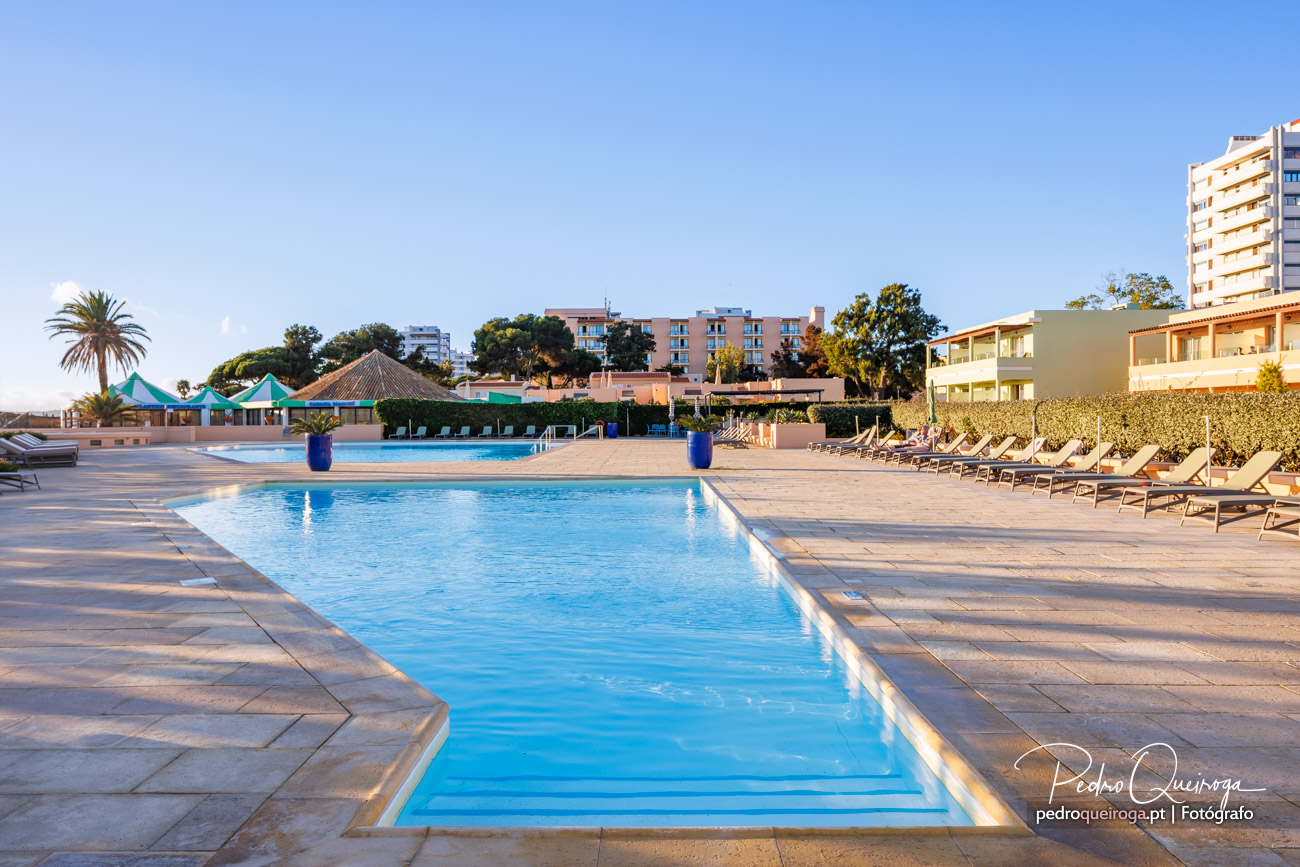 Piscinas amplas de resort ao entardecer, rodeadas por espreguiçadeiras e arquitetura moderna com edifícios ao fundo.