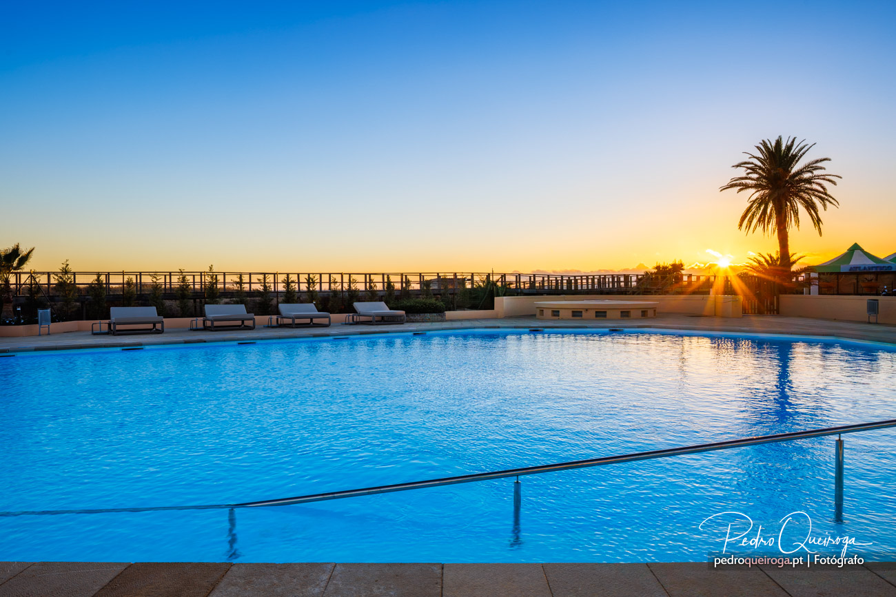 Piscina ampla ao pôr do sol com palmeira e espreguiçadeiras brancas junto ao horizonte dourado