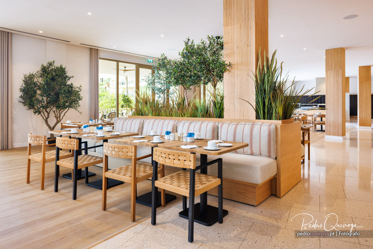 Restaurante luminoso com bancos estofados, mesas em madeira, cadeiras entrelaçadas e vegetação decorativa interior
