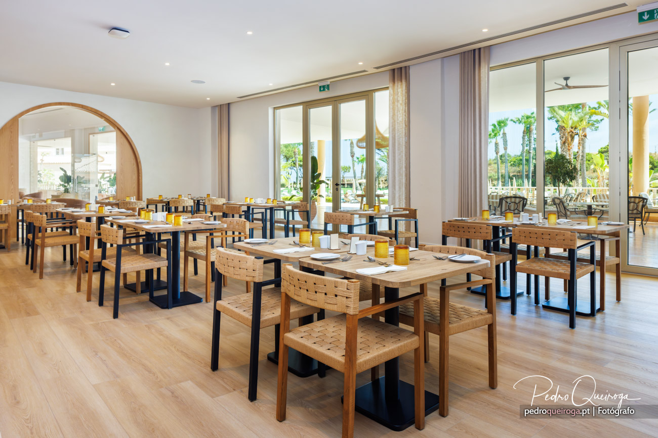 Sala de refeições luminosa com mesas em madeira e cadeiras entrelaçadas, amplas janelas e vista para palmeiras.