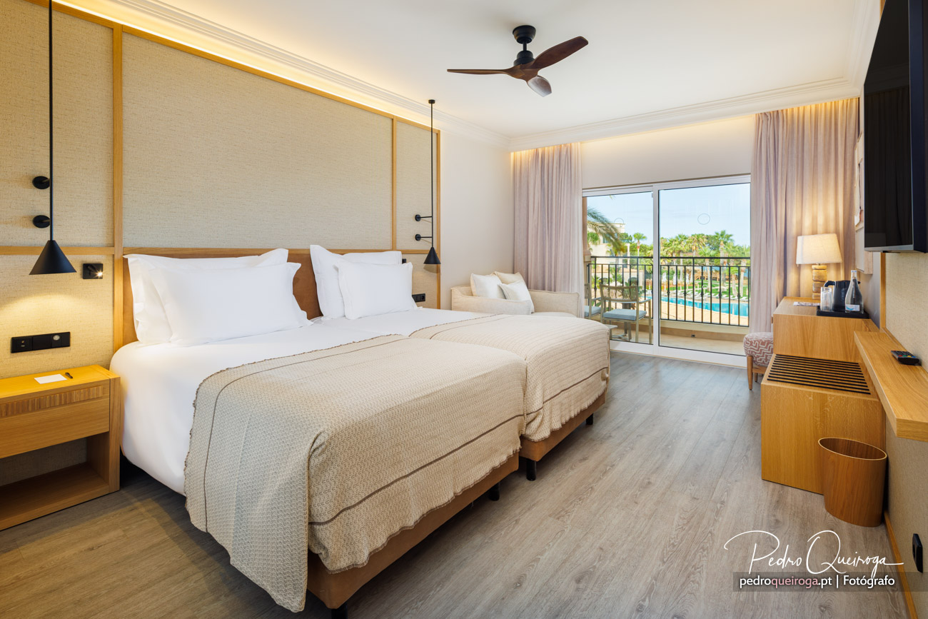 Quarto de hotel espaçoso com duas camas, varanda com vista para piscina e palmeiras, decoração em tons neutros e madeira.