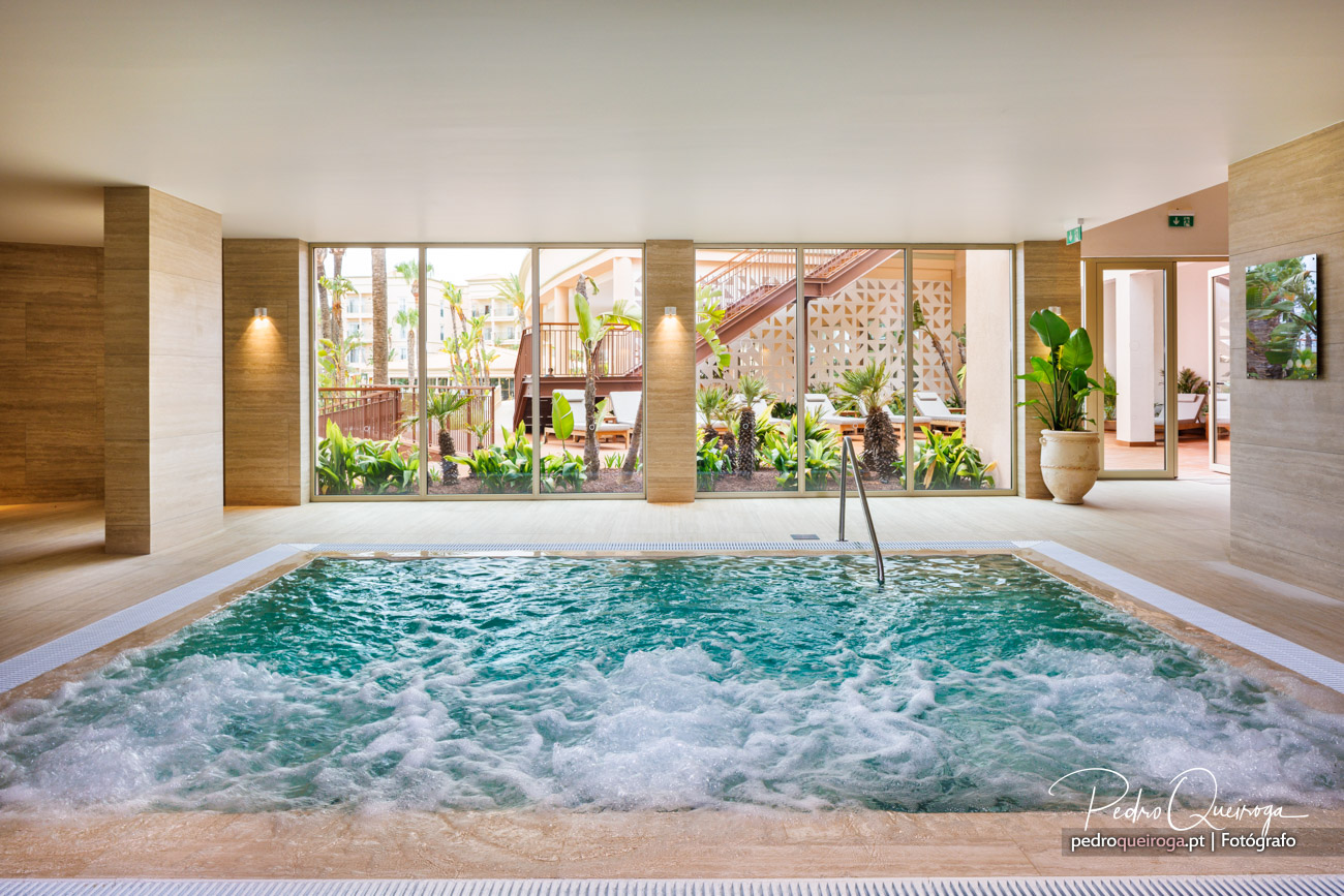 Jacuzzi interior com água em movimento, revestimentos em madeira e grandes janelas com vista para jardim tropical.