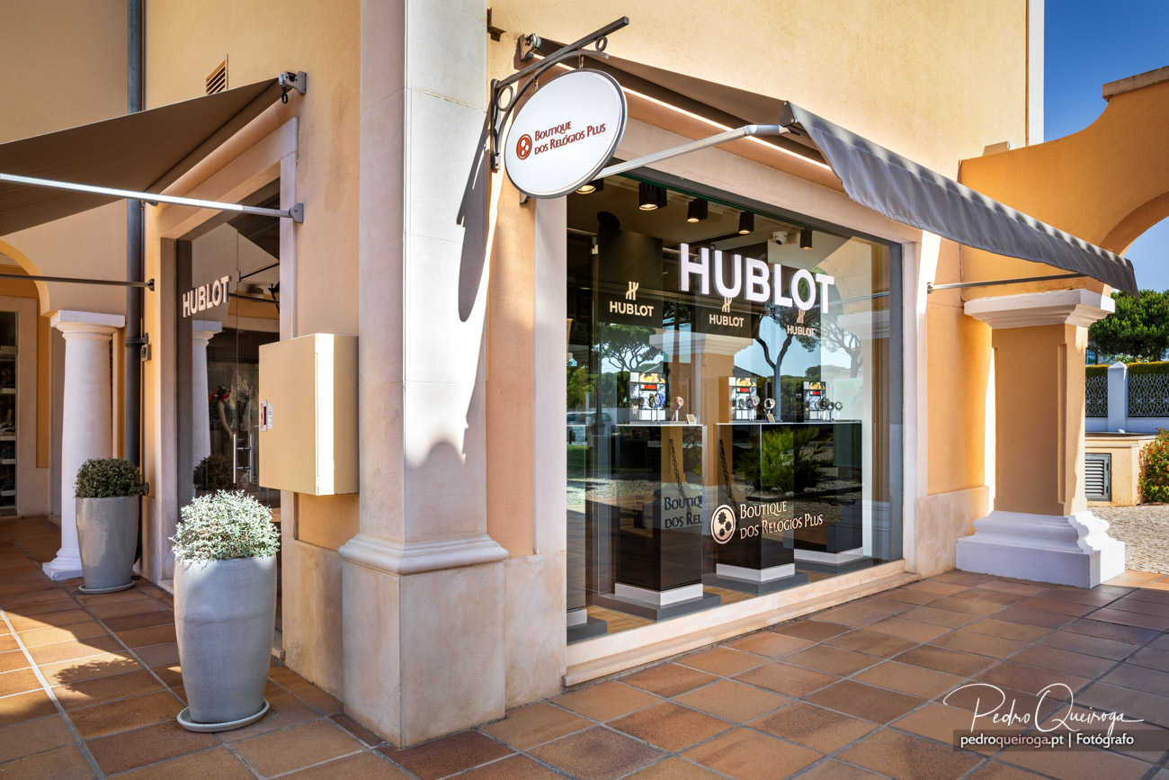 Fotografia de arquitetura da boutique Hublot com fachada elegante, colunas e toldos bege sob céu azul