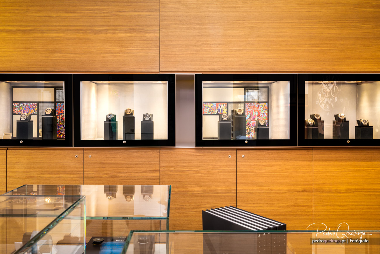 Montras elegantes de joalharia com relógios expostos em pedestais iluminados sobre painéis amadeirados e gavetas.