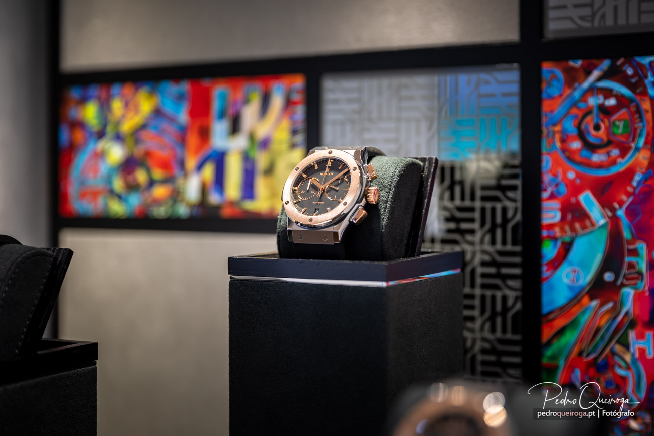 Relógio de luxo com caixa dourada e cinzenta sobre almofada preta, enquadrado por quadros artísticos coloridos ao fundo