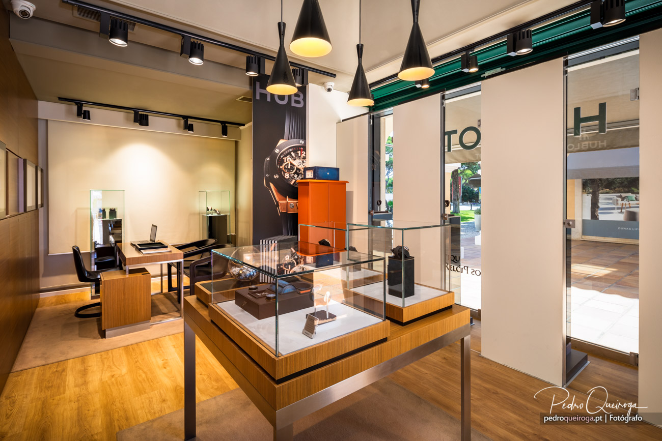 Interior moderno de loja de relógios com vitrines em vidro, iluminação pendente negra e móvel laranja ao fundo