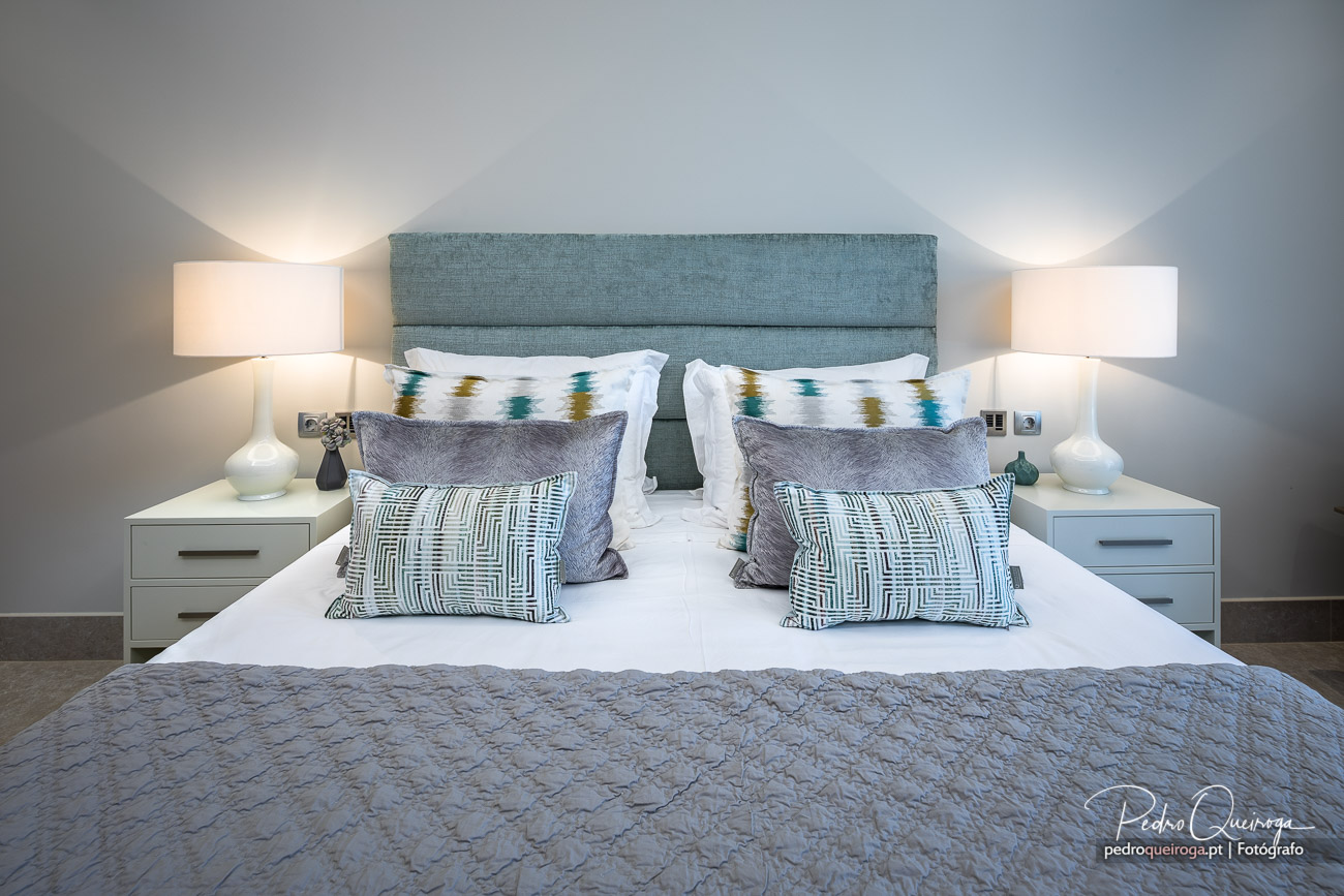 Quarto moderno com cabeceira azul estofada, almofadas decorativas e mesas de cabeceira brancas com candeeiros elegantes