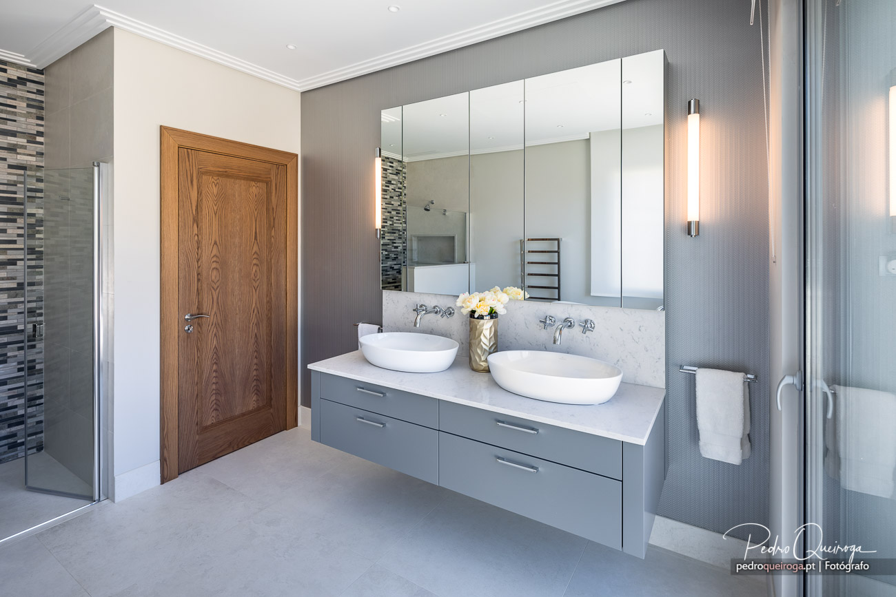 Casa de banho moderna com móvel cinza suspenso, duplo lavatório branco, espelho amplo e porta em madeira natural.