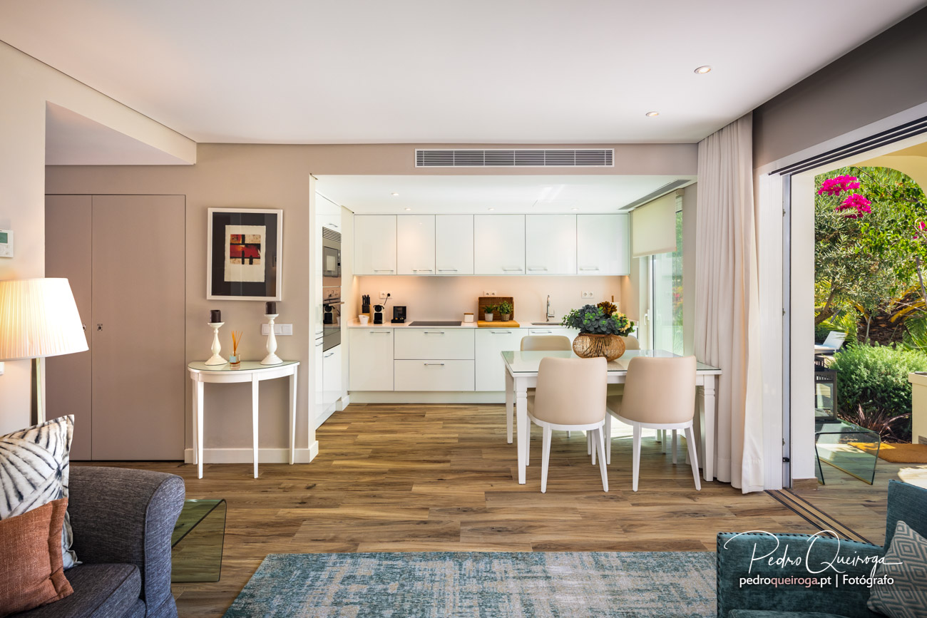 Espaço aberto com sala, cozinha branca moderna e zona de refeições, com vista para jardim verde e florido.